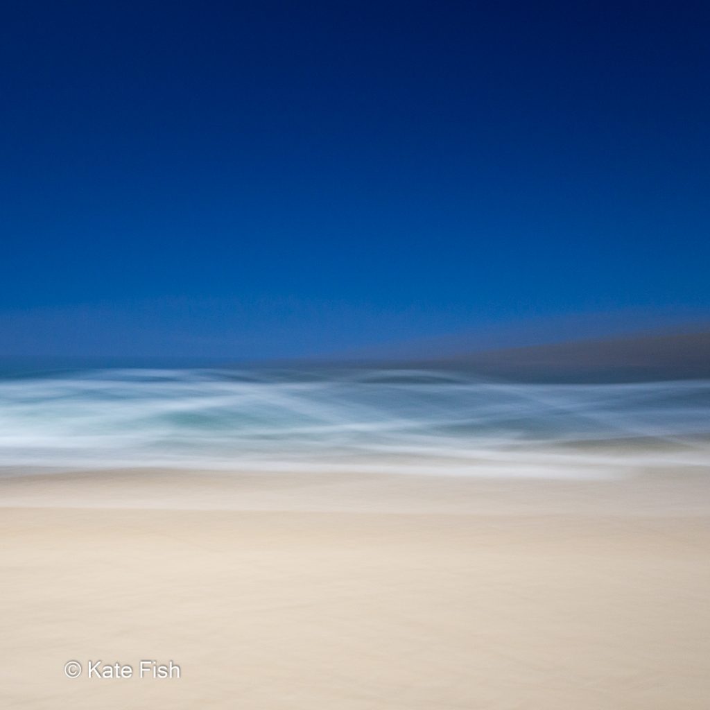 Bewußte Kamerabewegung (ICM)ICM Beach Art mit Wellenlinien aus den Schaumkronen der Wellen und beigem Sandstrand und tiefblauem Himmel, kreativ verwischt durch ziehen der Kamera während der Aufnahme