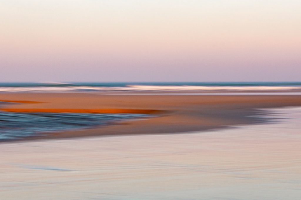 ICM Technik als Technik kreativ mit der Kamera zu sein, am Strand, durch einfaches Ziehen der Kamera verschwimmen hier die Konturen des Baches, des Sandstandes und der Wellen zu einem abstracten Bild mit geschwungenen Linienaus blau, orange und fast silber-rosa. Letzteres durch das Rosa am frühen Morgenhimmel über dem Strand von Mawgan Port in Cornwall
