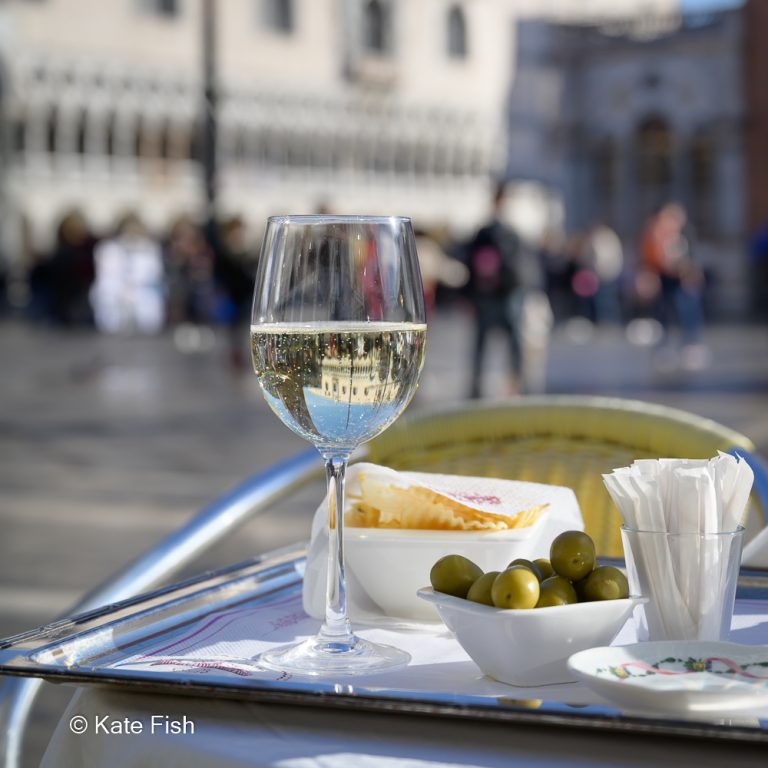 Spiegelung des Markus Doms im Sektglas auf einem Gedeck aus Oliven, Chips und Prosecco auf silbernem Tablet auf einem Tisch auf dem Markusplatz in Venedig als Beispiel, wie du kreativ mit der Kamera sein kannst