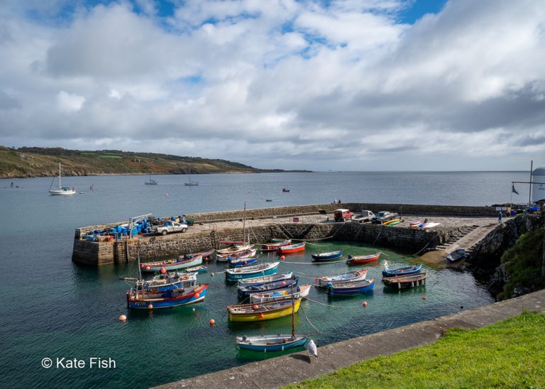 Coverack Hafen auf der Lizard Halbinsel in Cornwall mit kleinen Fischerbooten und einer Kaimauer, die den Hafen wie ein U umschließt. Türkisblaues Wasser, klar bis auf den Grund und im Hintergrund die Küste