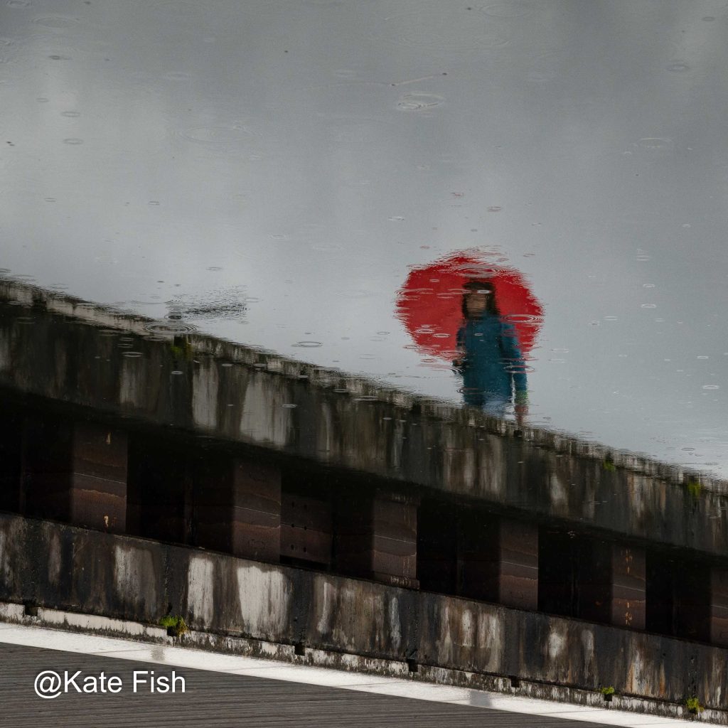 Mensch als Spiegelung im Wasser fotografieren. Hier im Regen mit rotem Regenschirm als Blickfang